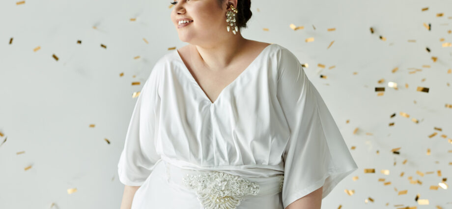 5 Tips for Wedding Dress Shopping Every Plus-Size Bride Should Follow 1 A plus-size bride smiling in her wedding dress as she stands in front of a white background with gold confetti falling around her.