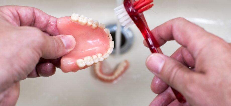 An individual using a small red toothbrush with red and white bristles to clean dentures over a sink.