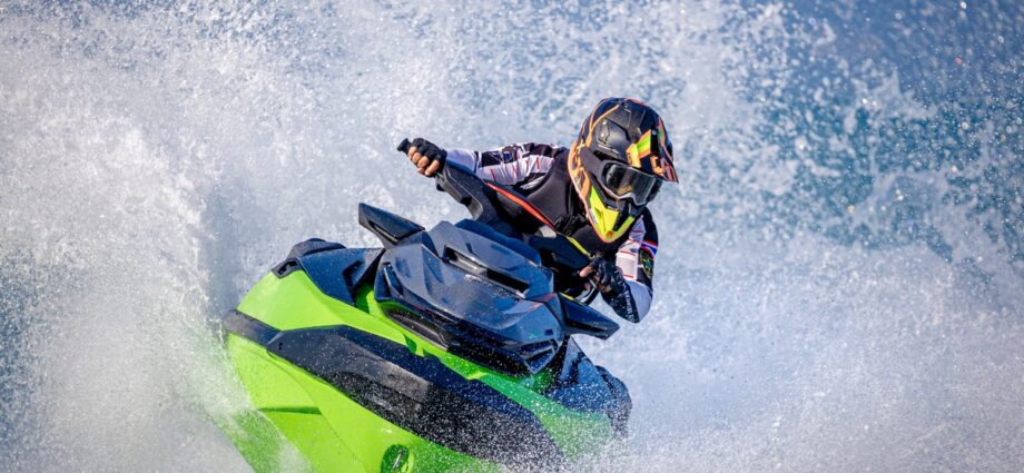 An individual in a helmet and goggles driving a black and green jet ski surrounded by splashing water.