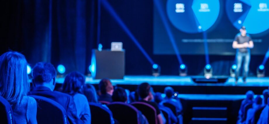 A large corporate event with blue stage lights and a presentation speaker on stage. There are people in the audience.