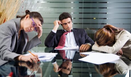 Three employees sit at a table. Two employees touch their heads and look frustrated. The other lays their head on the table.