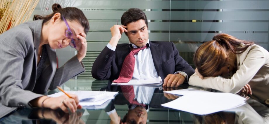 The Shocking Reasons Your Employees Don’t Click And How To Fix It Fast 1 Three employees sit at a table. Two employees touch their heads and look frustrated. The other lays their head on the table.