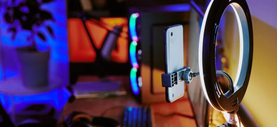 An online streaming studio setup. A gaming PC, keyboard, monitor, headphones, and a ring light sit on a wooden desk.