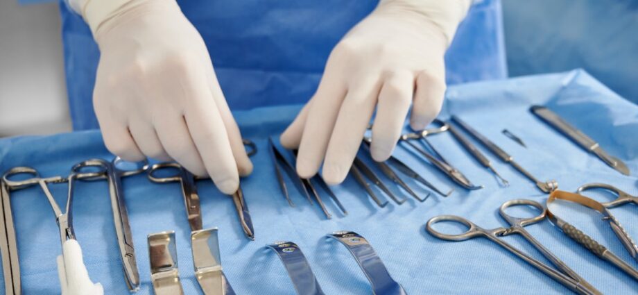 3 Most Important Medical Instruments to Know 1 A doctor wearing blue scrubs and white gloves stands in front of sterile surgical and medical instruments, ready for a procedure.