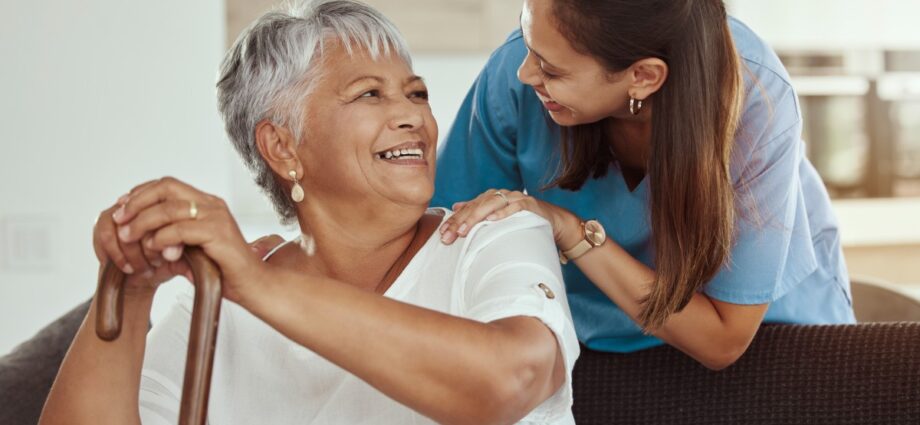 A caregiver offering at-home care to an old lady holding a cane while sitting down.
