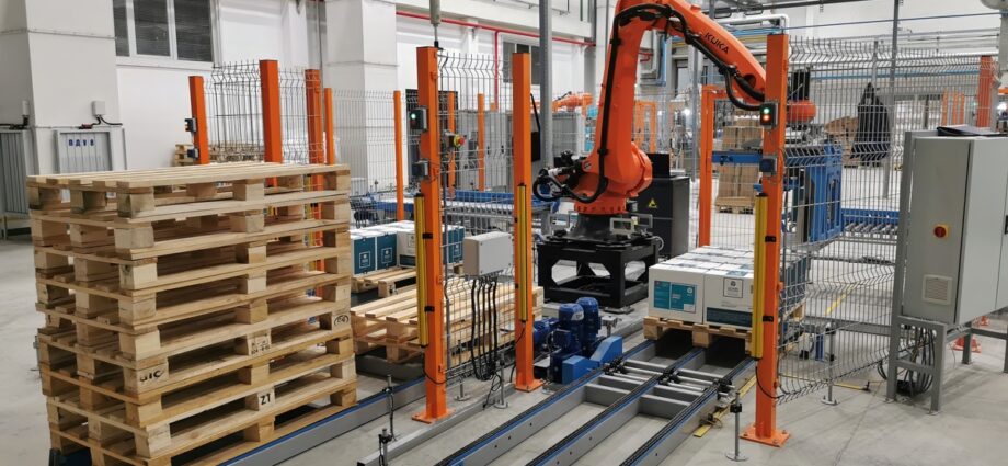 An automated palletizier system in a warehouse. There's a large, orange robotic arm and a stack of wooden pallets.