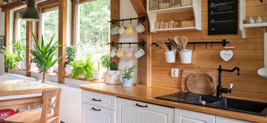 The interior of a rustic kitchen with white cabinets, wooden countertops, painted shelves, and hanging mugs.