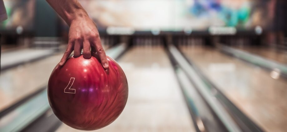 A close-up of a hand holding a red bowling ball with the upside-down outline of a seven on it in a bowling alley.