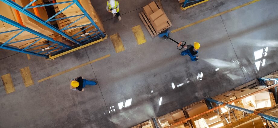 A top-down view of a business warehouse with different workers in yellow hard hats walking around and moving pallets.