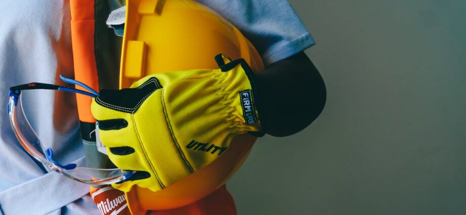 A person wearing construction work gear while holding a yellow hardhat with yellow gloves.