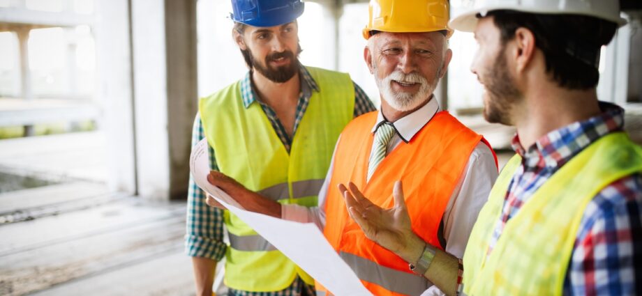 A construction manager smiles as he talks to two of his workers on a job site. He holds blueprints in his hand.
