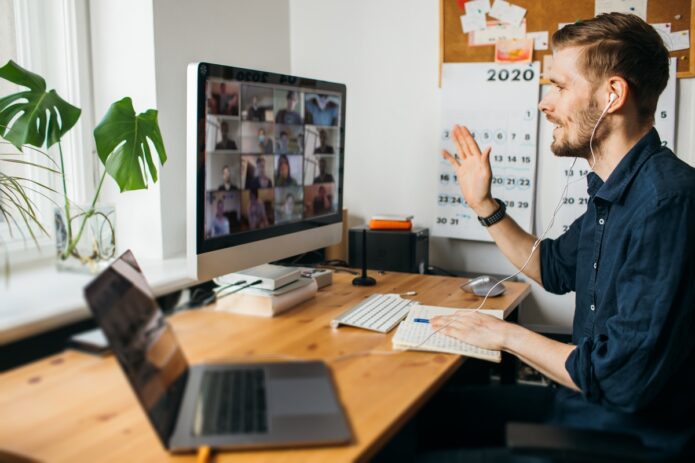 A young man working remotely at his home office on a zoom meeting with coworkers. He has headphones on and is taking notes.