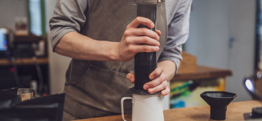 A barista wears a gray apron as they stand at the counter to prepare an AeroPress to brew fresh coffee.
