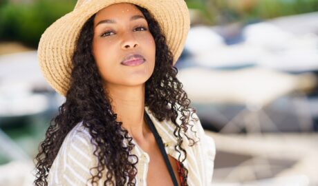 A woman with long curly hair wears a straw sun hat and light cover-up outfit standing near boats at a sunny marina.