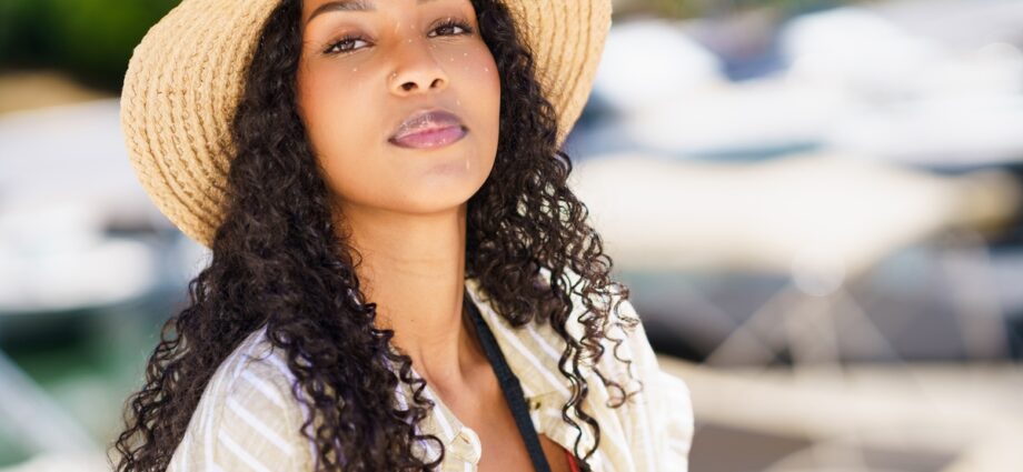 A woman with long curly hair wears a straw sun hat and light cover-up outfit standing near boats at a sunny marina.