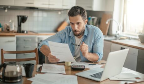 A man in a blue shirt sits at a table holding a document in front of an open laptop, coffee pot, and papers.