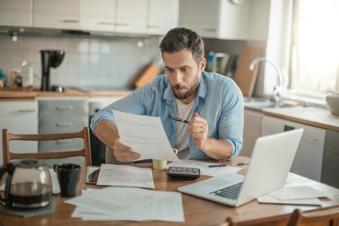 A man in a blue shirt sits at a table holding a document in front of an open laptop, coffee pot, and papers.