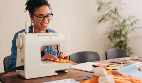 A smiling woman uses a sewing machine to stitch orange fabric at a worktable covered with textiles and other supplies.