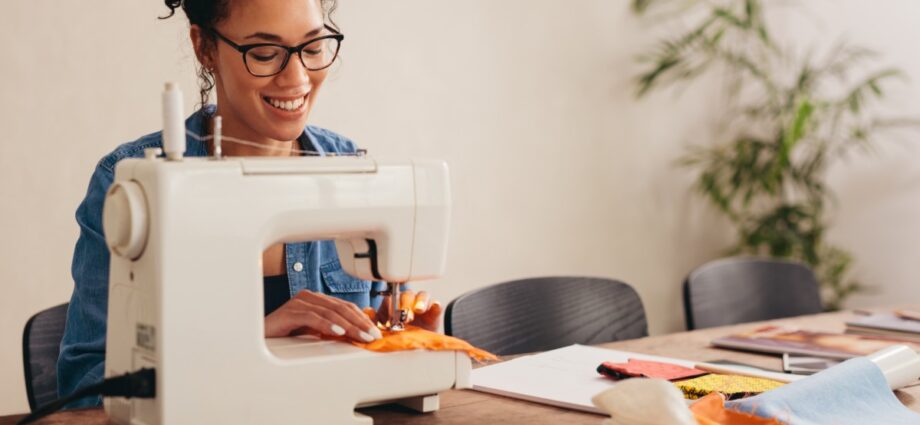 5 Easy Projects You Can Sew With No Experience 1 A smiling woman uses a sewing machine to stitch orange fabric at a worktable covered with textiles and other supplies.