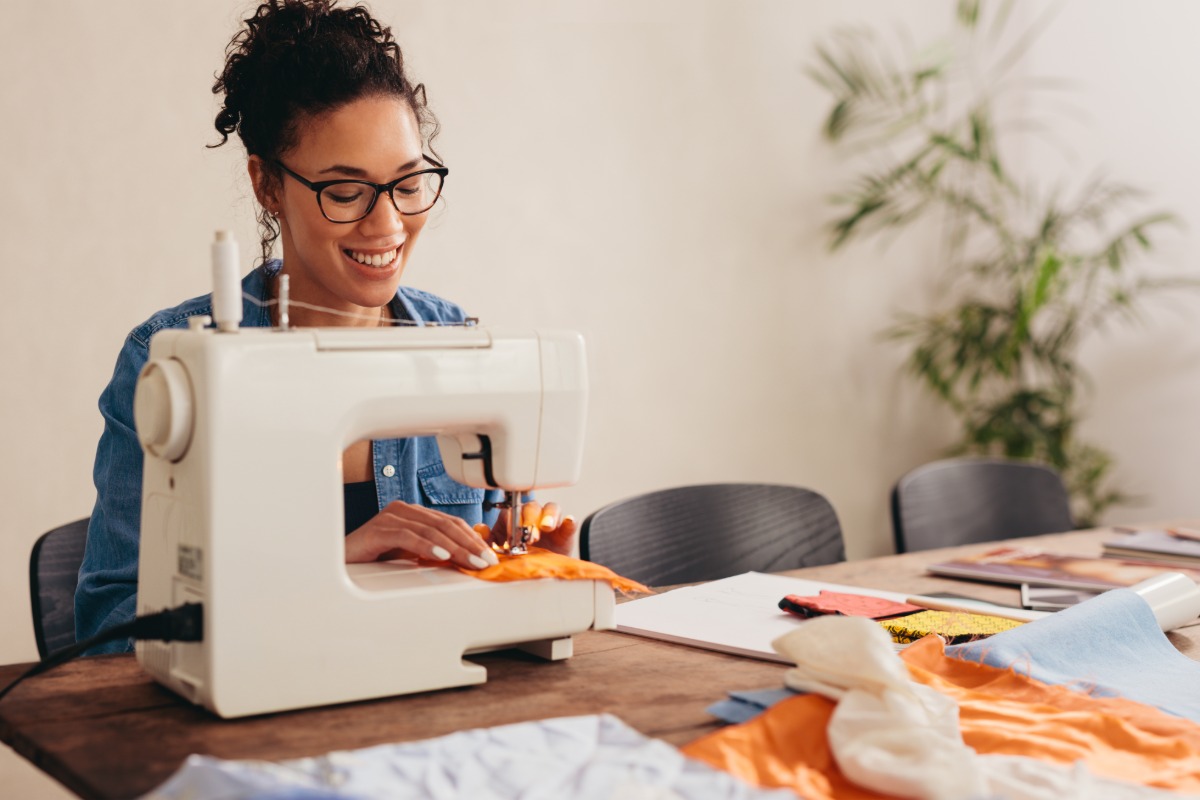 A smiling woman uses a sewing machine to stitch orange fabric at a worktable covered with textiles and other supplies.