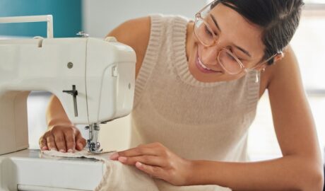 A young smiling woman in glasses runs a beige piece of fabric through a white sewing machine in a bright room.