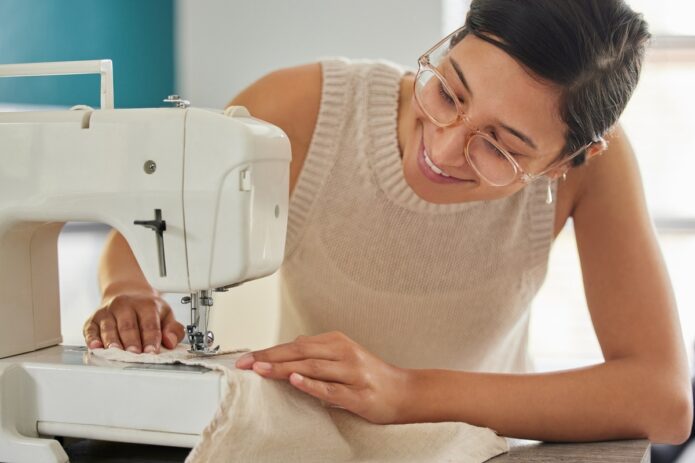 A young smiling woman in glasses runs a beige piece of fabric through a white sewing machine in a bright room.