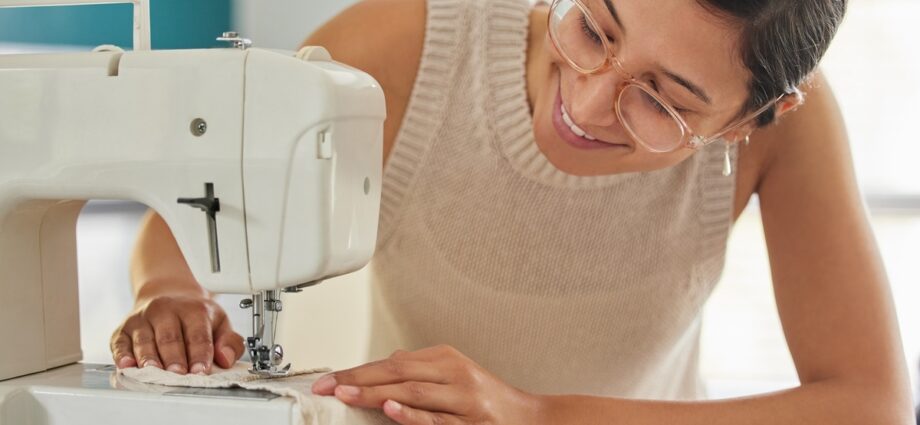 A young smiling woman in glasses runs a beige piece of fabric through a white sewing machine in a bright room.