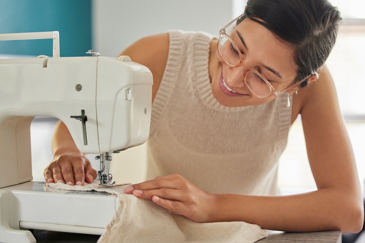A young smiling woman in glasses runs a beige piece of fabric through a white sewing machine in a bright room.