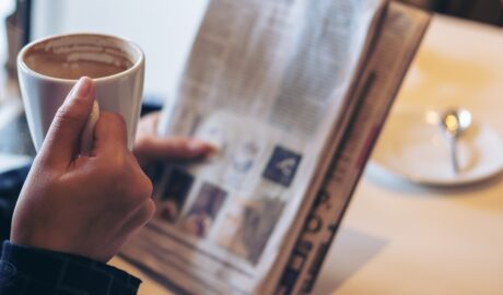 Someone reads the paper with their left hand and drinks out of a hot coffee mug with their right hand.
