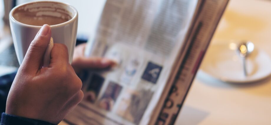 Someone reads the paper with their left hand and drinks out of a hot coffee mug with their right hand.