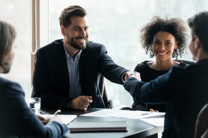 Improving Business Relationships Through Strict Standards 1 A smiling man in a light gray suit and glasses shaking hands with another person across a desk in an office.
