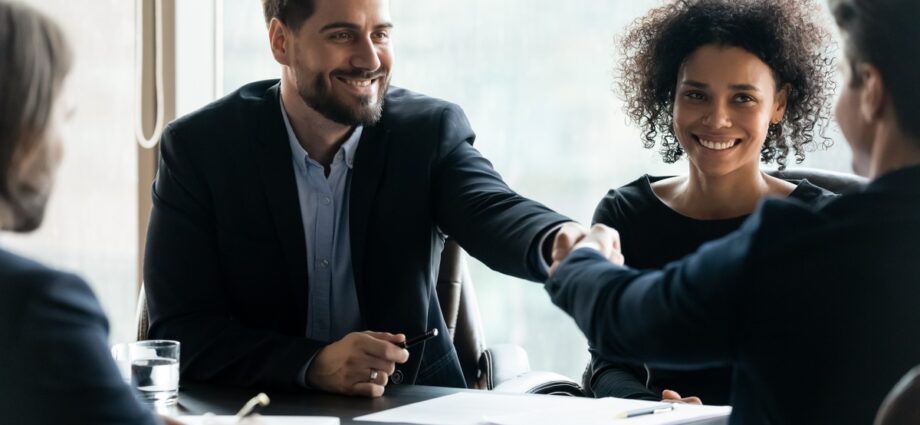 Improving Business Relationships Through Strict Standards 1 A smiling man in a light gray suit and glasses shaking hands with another person across a desk in an office.