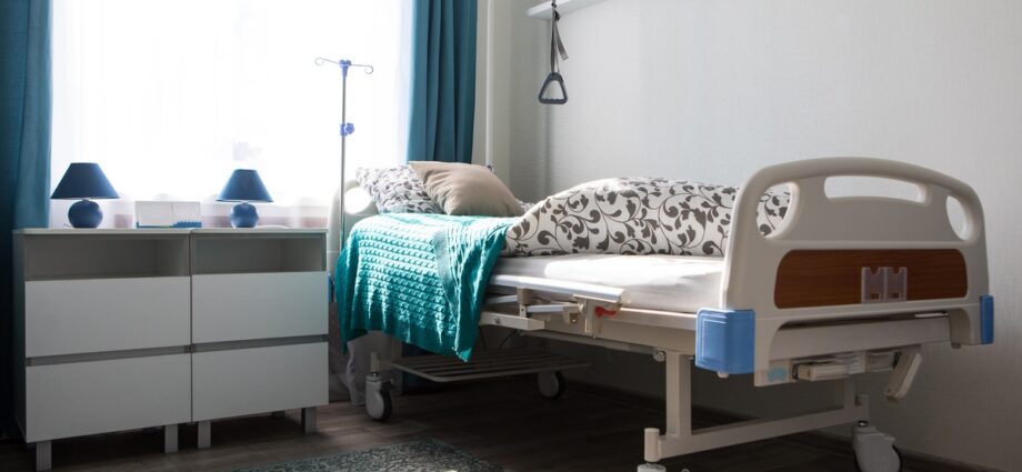 A hospital bed tucked away into the corner of a bedroom. The bed has a floral comforter and a blue knit blanket.