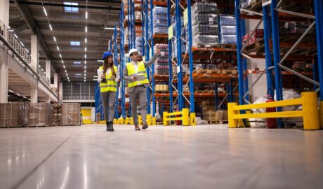 Two managers wearing hard hats and safety vests walking through a warehouse aisle lined with pallet racks.