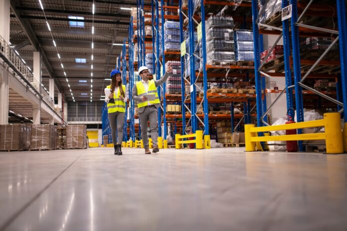 Two managers wearing hard hats and safety vests walking through a warehouse aisle lined with pallet racks.