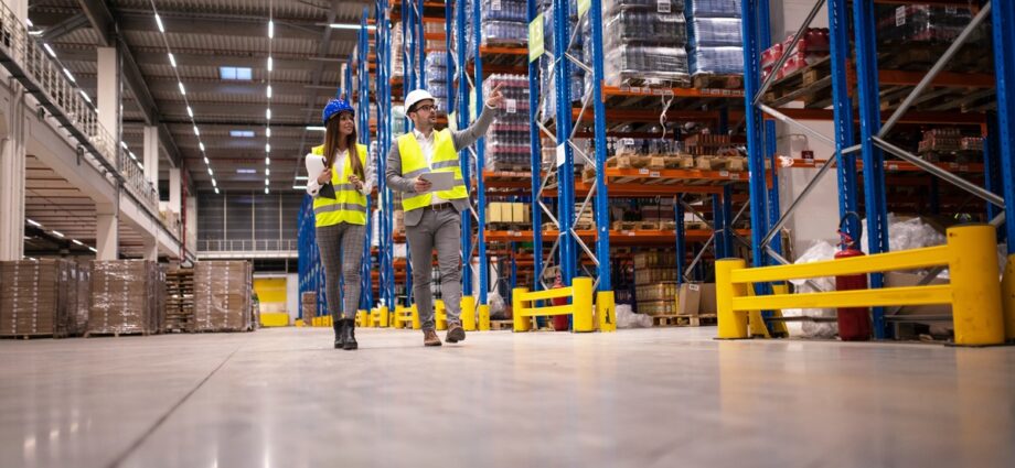 Two managers wearing hard hats and safety vests walking through a warehouse aisle lined with pallet racks.