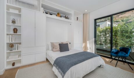 A white bedroom with a bed under built-in shelves and cabinets, a blue chair, rug, and large glass doors.