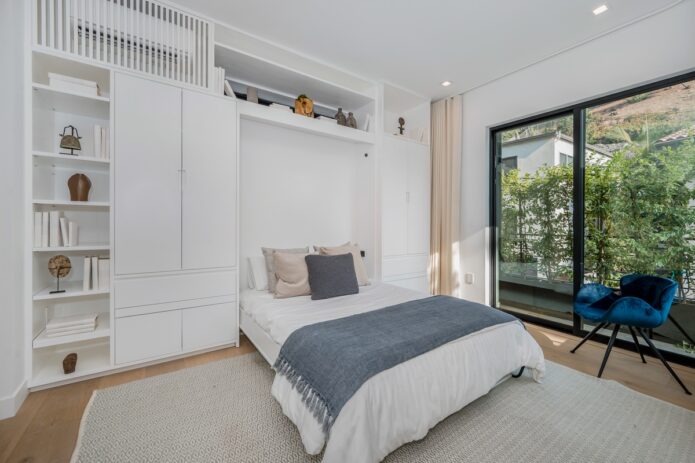 A white bedroom with a bed under built-in shelves and cabinets, a blue chair, rug, and large glass doors.