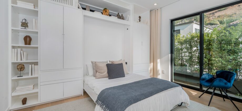 How Flexible Furniture Adapts to Changing Household Needs 1 A white bedroom with a bed under built-in shelves and cabinets, a blue chair, rug, and large glass doors.
