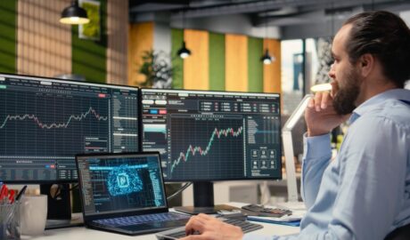 A man in a dress shirt sits at a desk with multiple monitors, a laptop, and a keyboard with various economic graphs.