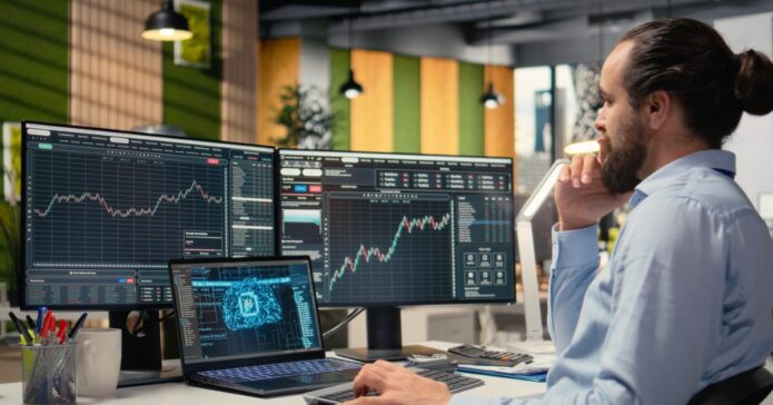 10 Ways How Traders Use Technical Analysis Tools 1 A man in a dress shirt sits at a desk with multiple monitors, a laptop, and a keyboard with various economic graphs.