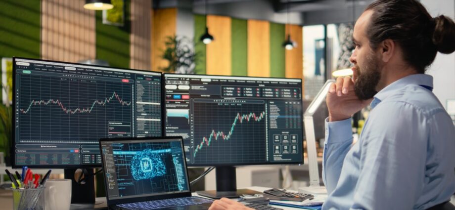 10 Ways How Traders Use Technical Analysis Tools 1 A man in a dress shirt sits at a desk with multiple monitors, a laptop, and a keyboard with various economic graphs.