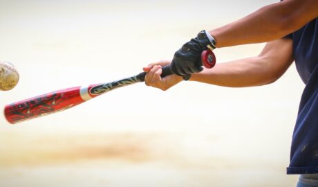 A close-up of a bat being swung by a young player with one batting glove and shorts at a ball in mid-flight.
