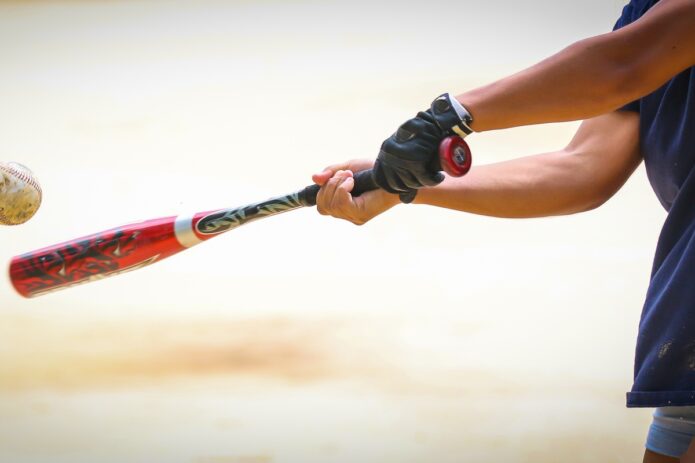 A close-up of a bat being swung by a young player with one batting glove and shorts at a ball in mid-flight.