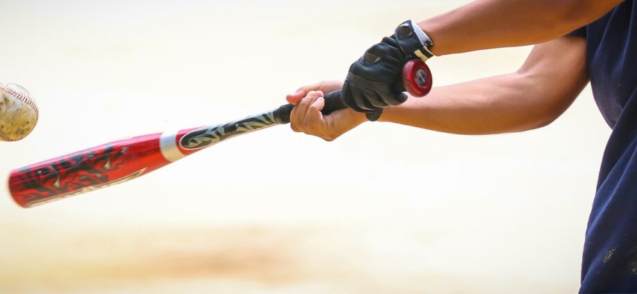 A close-up of a bat being swung by a young player with one batting glove and shorts at a ball in mid-flight.